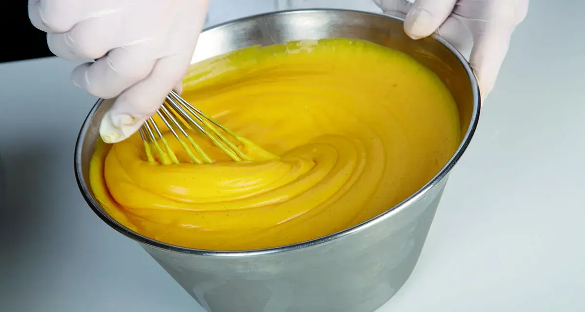 Mani guantate montano una crema pasticcera perfetta, come insegna Iginio Massari. La consistenza è vellutata e il colore giallo intenso.