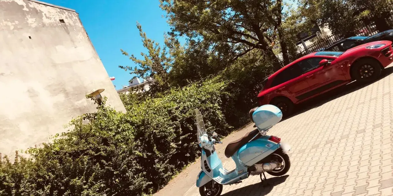 Błękitny skuter czeka na przejażdżkę. W tle czerwony samochód i zielone drzewa. Idealne miejsce na parking moto.