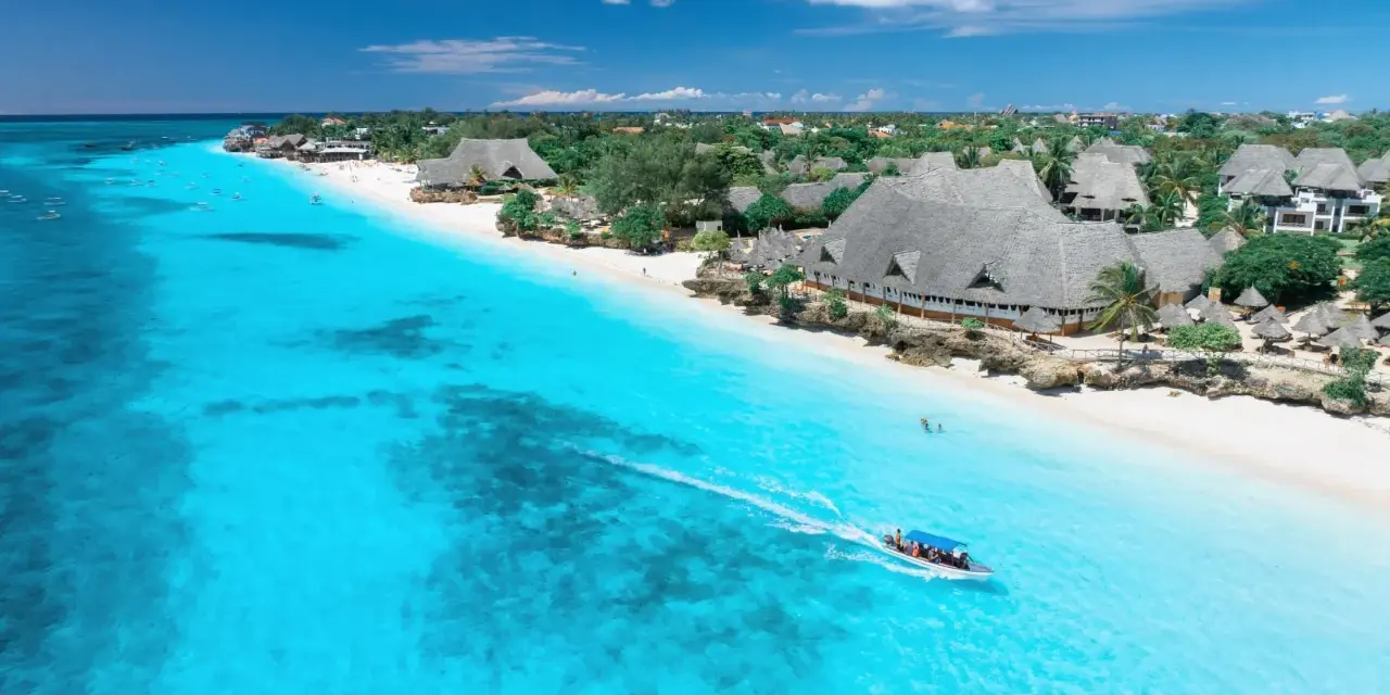 Widok na turkusowe morze i białą plażę z hotelami, kiedy na Zanzibarze. Łódź motorowa z pasażerami płynie po wodzie.