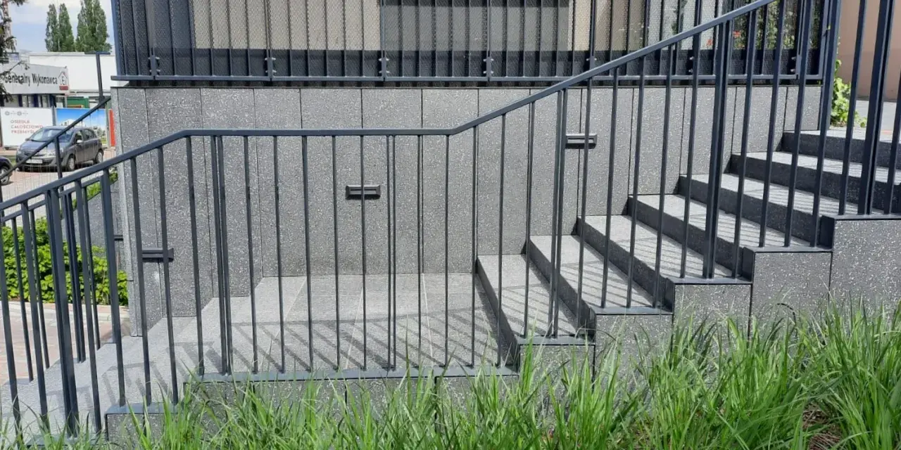 Nowoczesne schody zewnętrzne z metalową balustradą prowadzą do budynku. Wokół bujna zieleń.