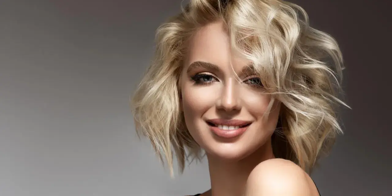 Mujer sonriente con cabello rubio ondulado y cara alargada.
