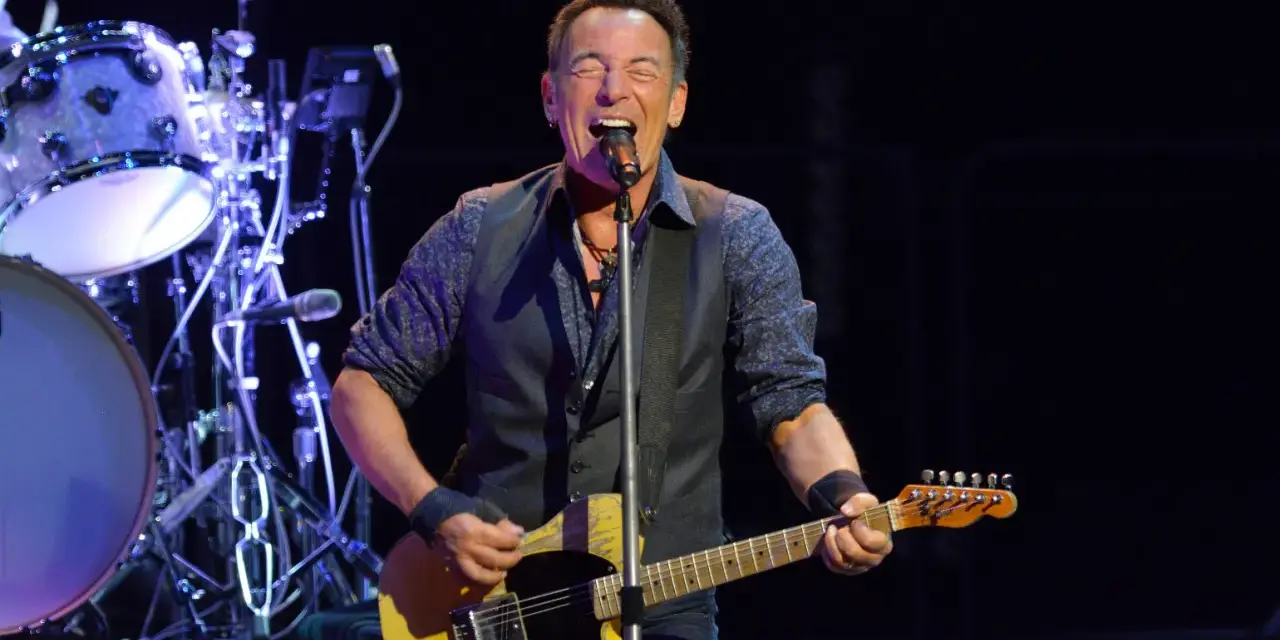 Bruce Springsteen singt und spielt seine ikonische Bruce Springsteen Gitarre auf der Bühne, während die Band im Hintergrund spielt.