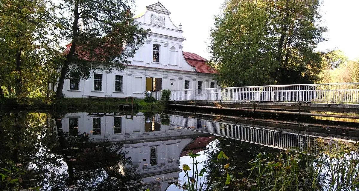 Biały kościół na wodzie w Zwierzyńcu, z odbiciem w tafli jeziora i mostkiem.