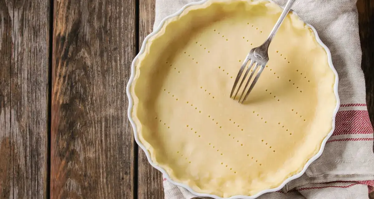 Pâte brisée sans beurre dans un plat à tarte, prête à être garnie. Une fourchette est plantée dans la pâte.