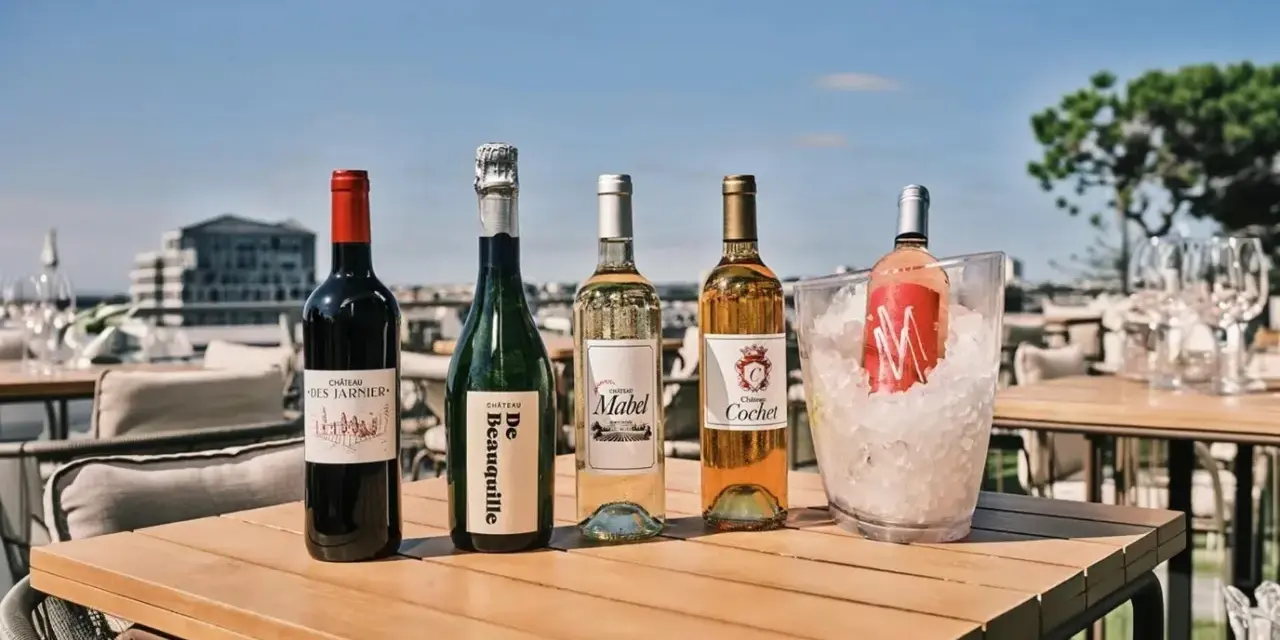Pięć butelek wina, w tym jedno bordeaux, na drewnianym stole na tarasie z widokiem na miasto.