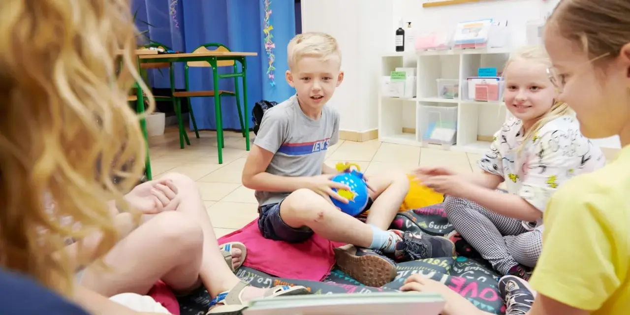 Chłopiec, około 10 lat, siedzi na podłodze z niebieską zabawką. Wokół niego inne dzieci.