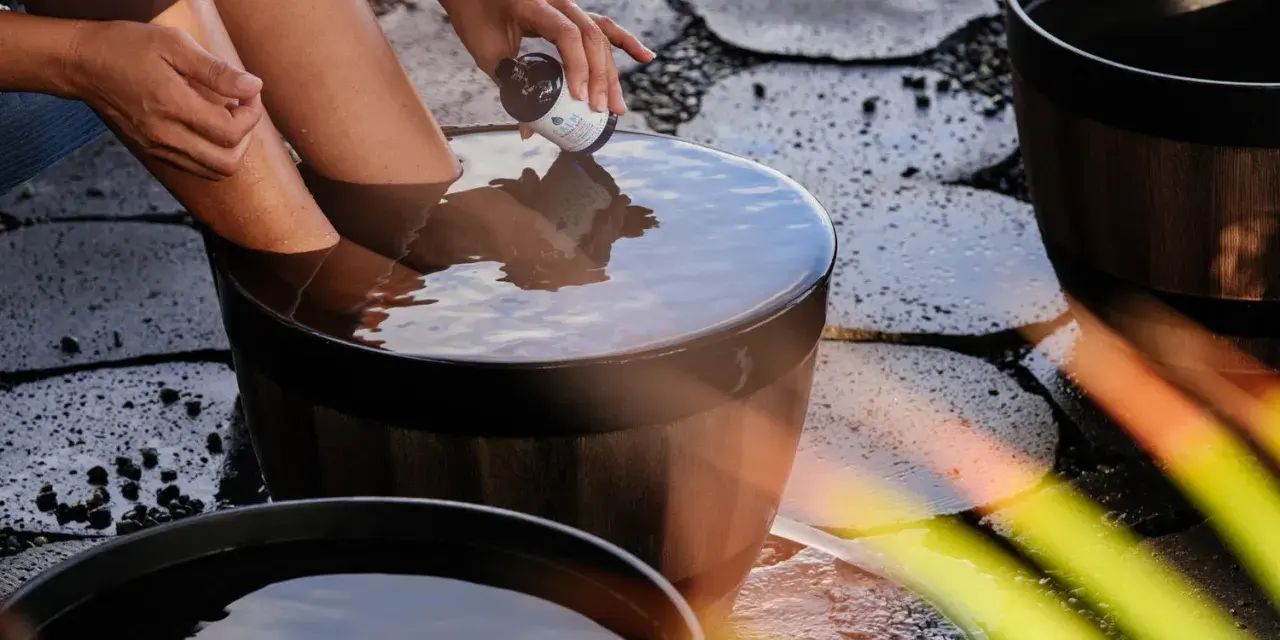 A person adds a few drops from a small bottle to a wooden tub filled with water, creating the best natural foot soak.