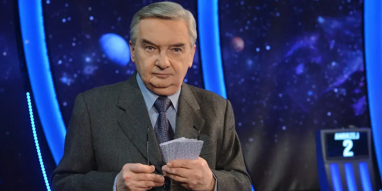 Starsza osoba w garniturze trzyma karty z napisem "1 z 10", gotowa do emisji.