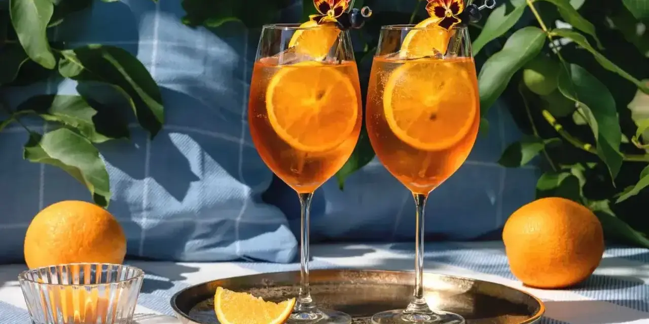 Dwa kieliszki Aperol Spritz z plastrami pomarańczy i kwiatami. Czy aperol można pić sam? Tak, ale najlepiej smakuje w towarzystwie!