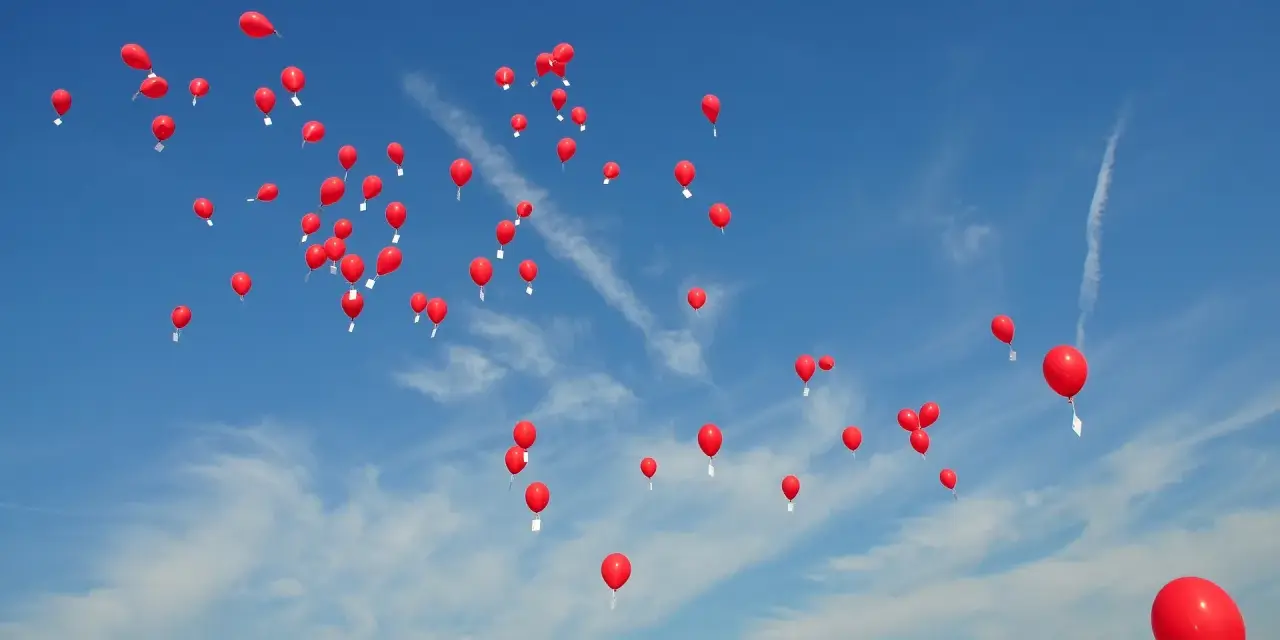 Många röda heliumballonger stiger mot en klarblå himmel med tunna moln.