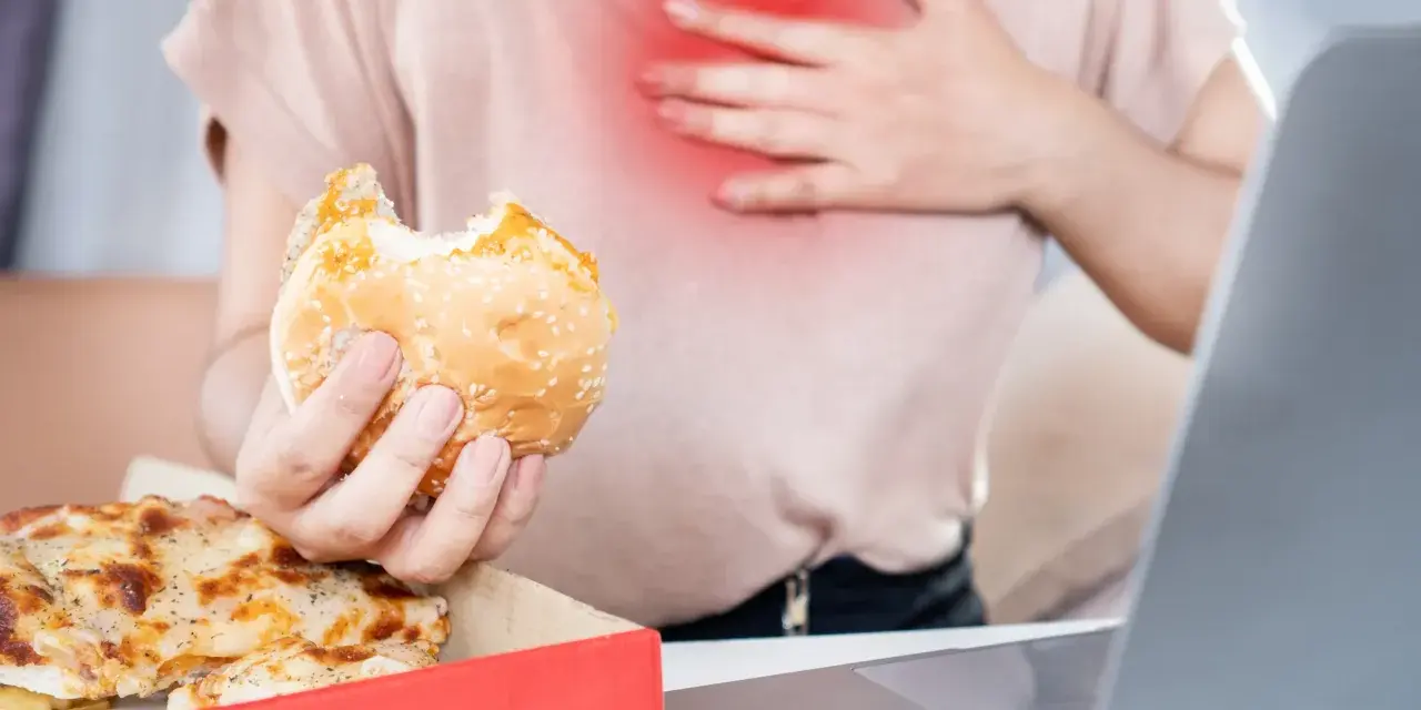 Kobieta z bólem w klatce piersiowej trzyma hamburgera. Zastanawia się, co pić na zgagę po zjedzeniu pizzy i fast foodów.