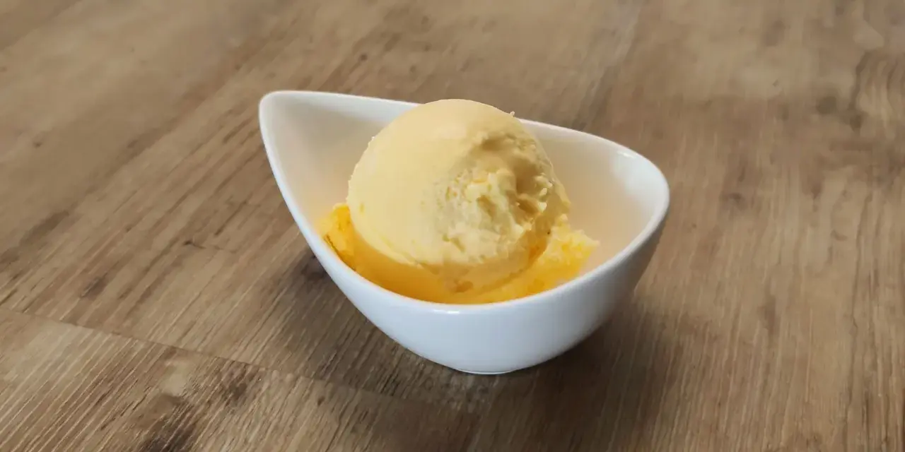 Ein Löffel cremiges Mandarinen Eis in einer weißen Schale auf Holzhintergrund.
