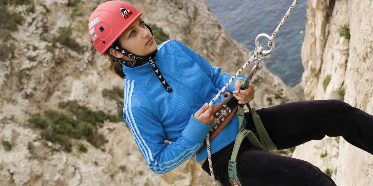 Junge Frau beim Abseilen an einer Felswand, Malta Klettern. Sie trägt einen roten Helm und eine blaue Jacke.