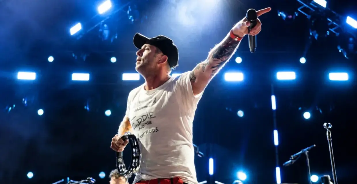 Wokalista z tatuażami i czapką na głowie prowadzi koncert One Republic, wskazując ręką w górę i trzymając tamburyn.