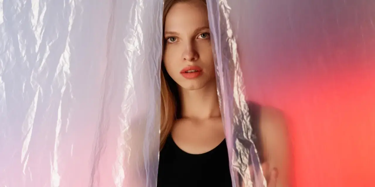 A young woman peeks through crinkled plastic sheet materials, her face illuminated by soft, colorful light.