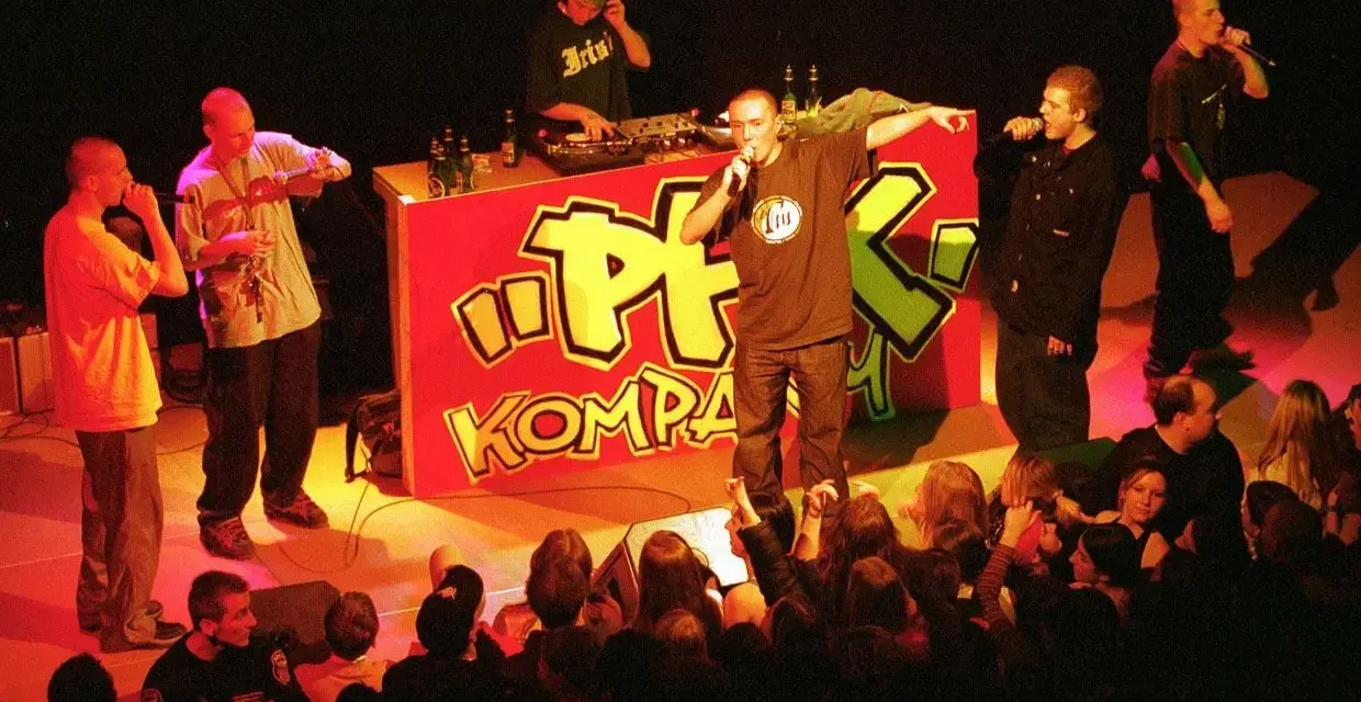Raperzy na scenie przed czerwonym banerem z napisem "PFK Kompakt", publiczność w pierwszym rzędzie.
