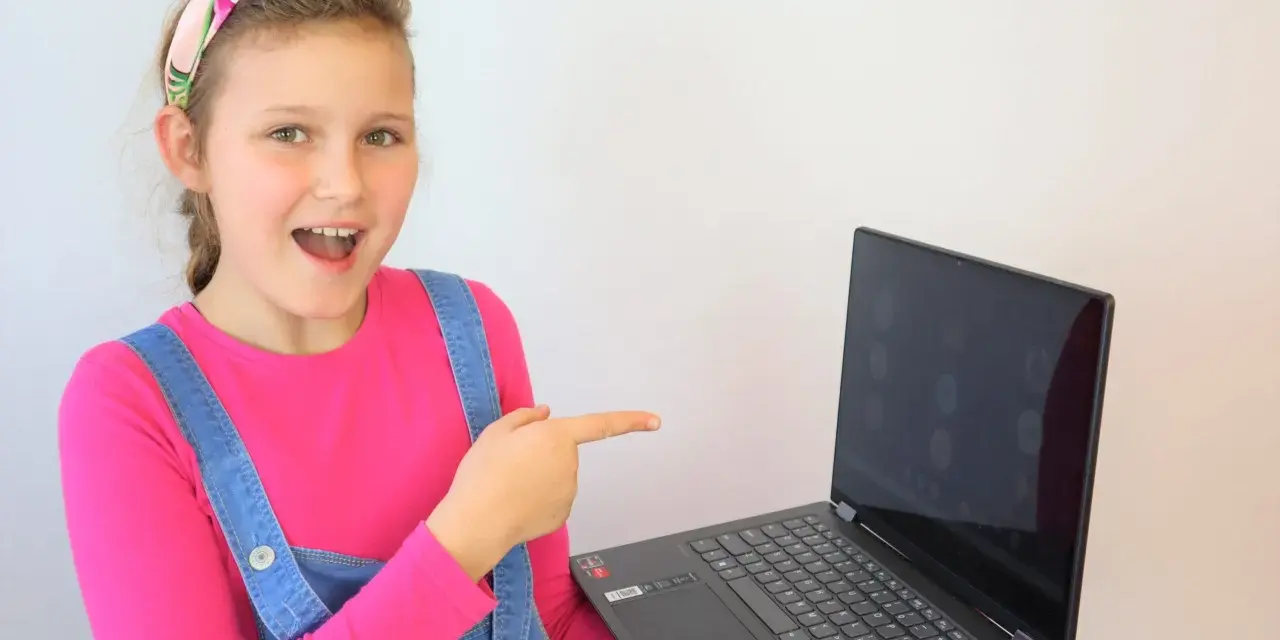 Uśmiechnięta dziewczynka w ogrodniczkach wskazuje na ekran laptopa, gdzie widać okienka z ikonami. Idealne miejsce na naukę angielskiego online dla dzieci.