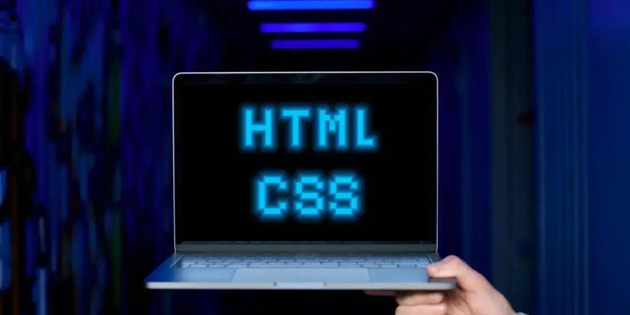 <p>Dłoń trzyma laptopa, na ekranie którego świeci napis "HTML CSS" w niebieskich pikselach. Tło jest ciemne, z niebieskimi neonami.</p>