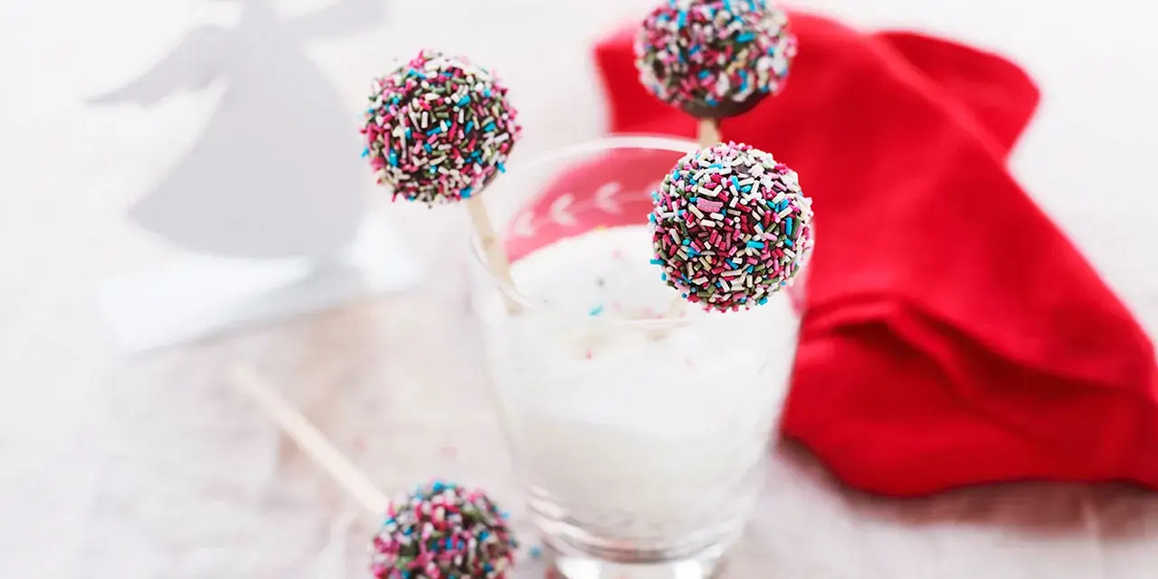 Söta cake pops med pepparkakssmak, dekorerade med färgglada strössel. En vit ängel i bakgrunden.