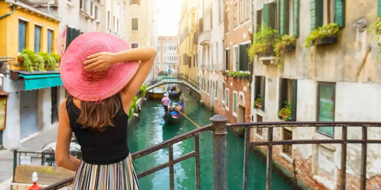 Donna con cappello rosa ammira un canale a Venezia, con gondole e palazzi storici. Un'esperienza da vivere a piedi, tra Santa Lucia e Piazza San Marco.