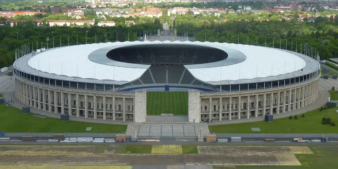 Das Olympiastadion Berlin mit seinem markanten Dach und der Skyline im Hintergrund. In der Ferne ragt der Fernsehturm, der wie ein glockenturm Berlin überragt.