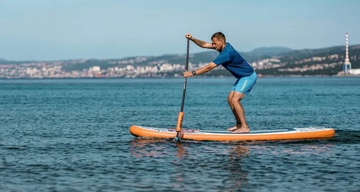 Mężczyzna na desce sup wiosłuje po spokojnym morzu, w tle widać miasto.