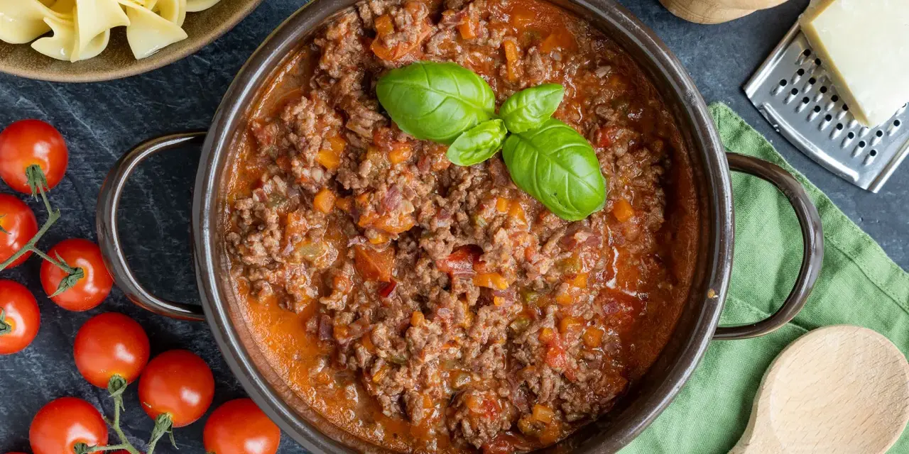 Herzhaftes Ragout Bolognese in einem Topf, garniert mit Basilikum. Daneben frische Tomaten, Bandnudeln und Parmesan.