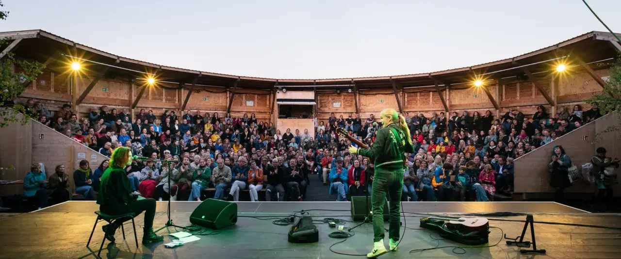 Konzert auf dem Tempelhofer Feld: Eine Musikerin spielt Gitarre vor einem begeisterten Publikum in einem hölzernen Amphitheater. Ein wahrer luftschloss tempelhofer feld Traum.