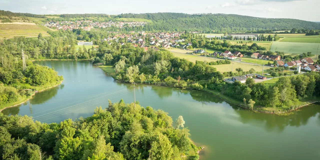 Grüne Hügel, ein Dorf und der glitzernde Klostersee Triefenstein unter blauem Himmel.
