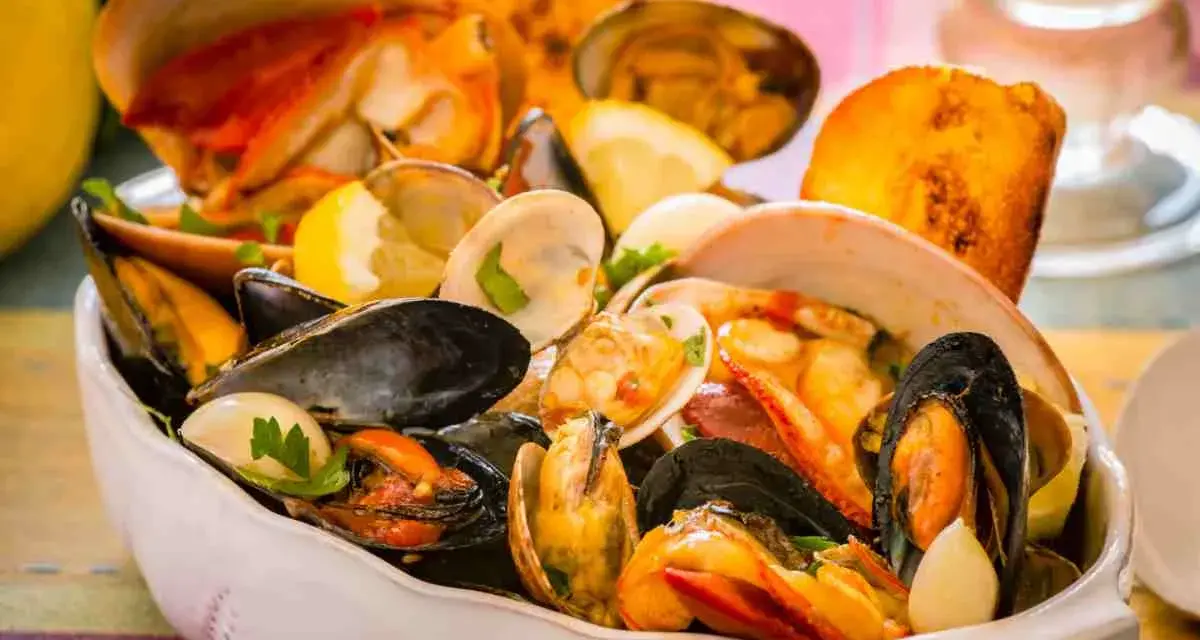 Un fumetto di pesce ricco di cozze, vongole e crostacei, guarnito con limone e prezzemolo, servito in una ciotola bianca.