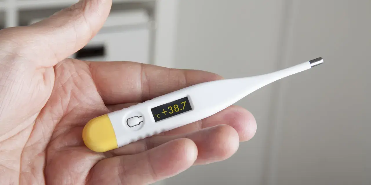 Dłoń trzyma termometr wskazujący 38.7°C. Czas na działanie, jak zbić gorączkę.