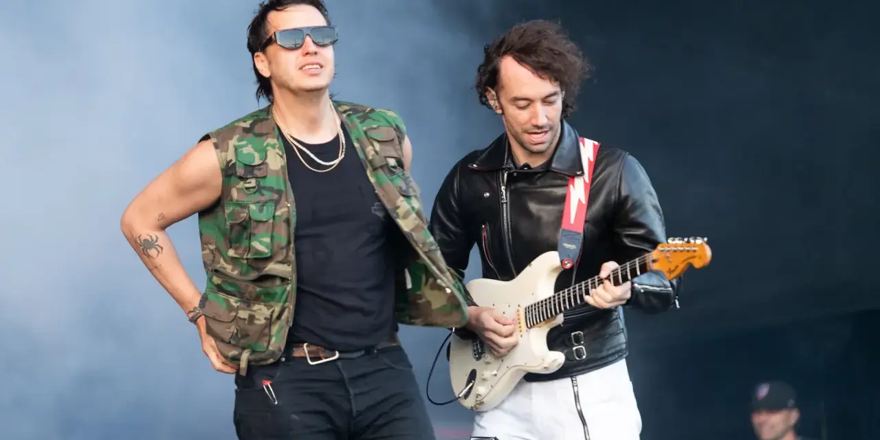 Albert Hammond Jr. w kamizelce moro i okularach stoi obok gitarzysty w skórzanej kurtce, grającego na białej gitarze.