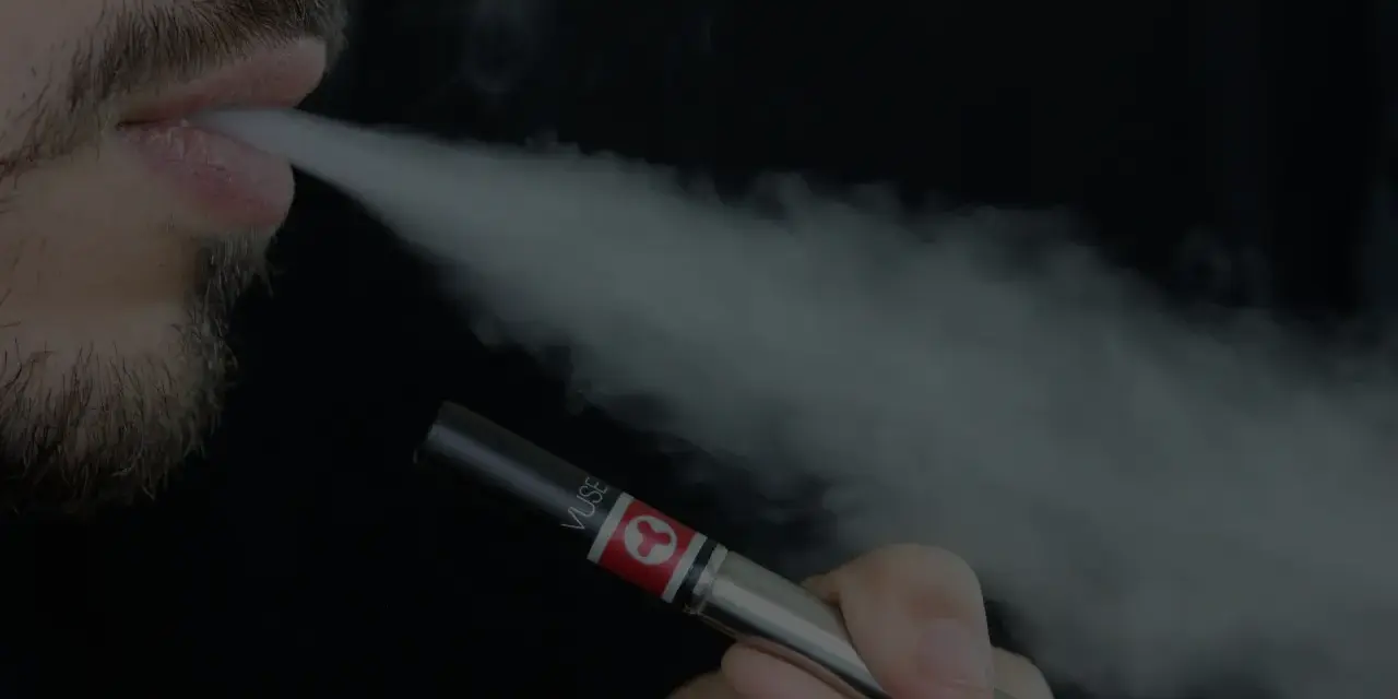 Un hombre con barba exhala vapor de un vapeador, similar al estilo Heisenberg.