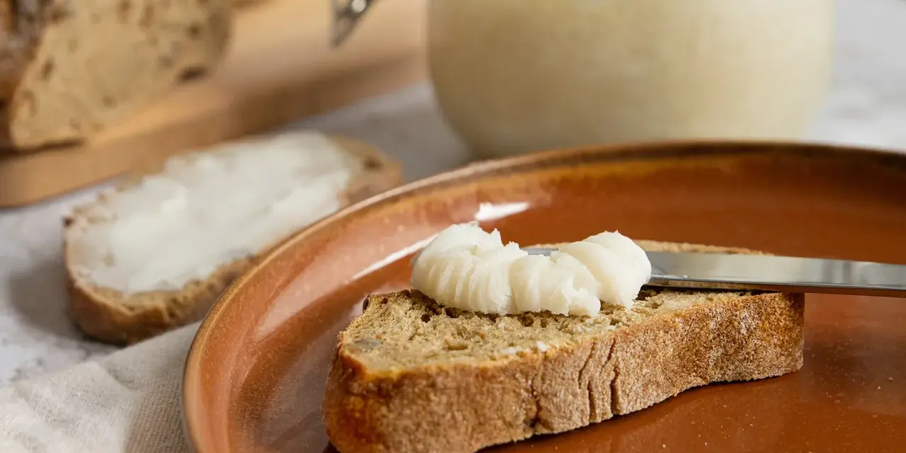 Ein Glas mit selbstgemachtem Schmalz steht bereit. Eine Scheibe Brot wird gerade mit dem Schmalz bestrichen, ein Genuss für jeden, der Schmalz selber machen möchte.