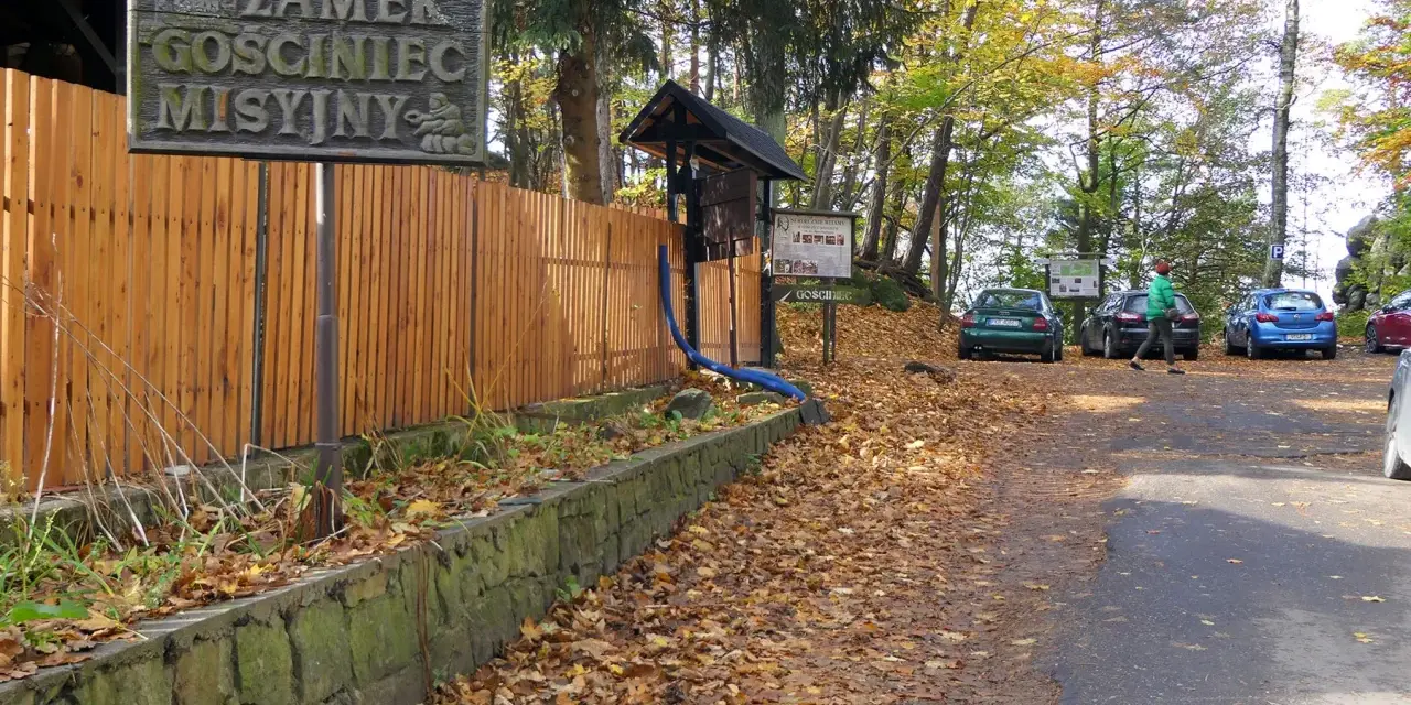 Tablica z napisem "Zamek GOSCINIEC MISYJNY" przy drewnianym płocie. Jesienny parking z samochodami i opadłymi liśćmi.