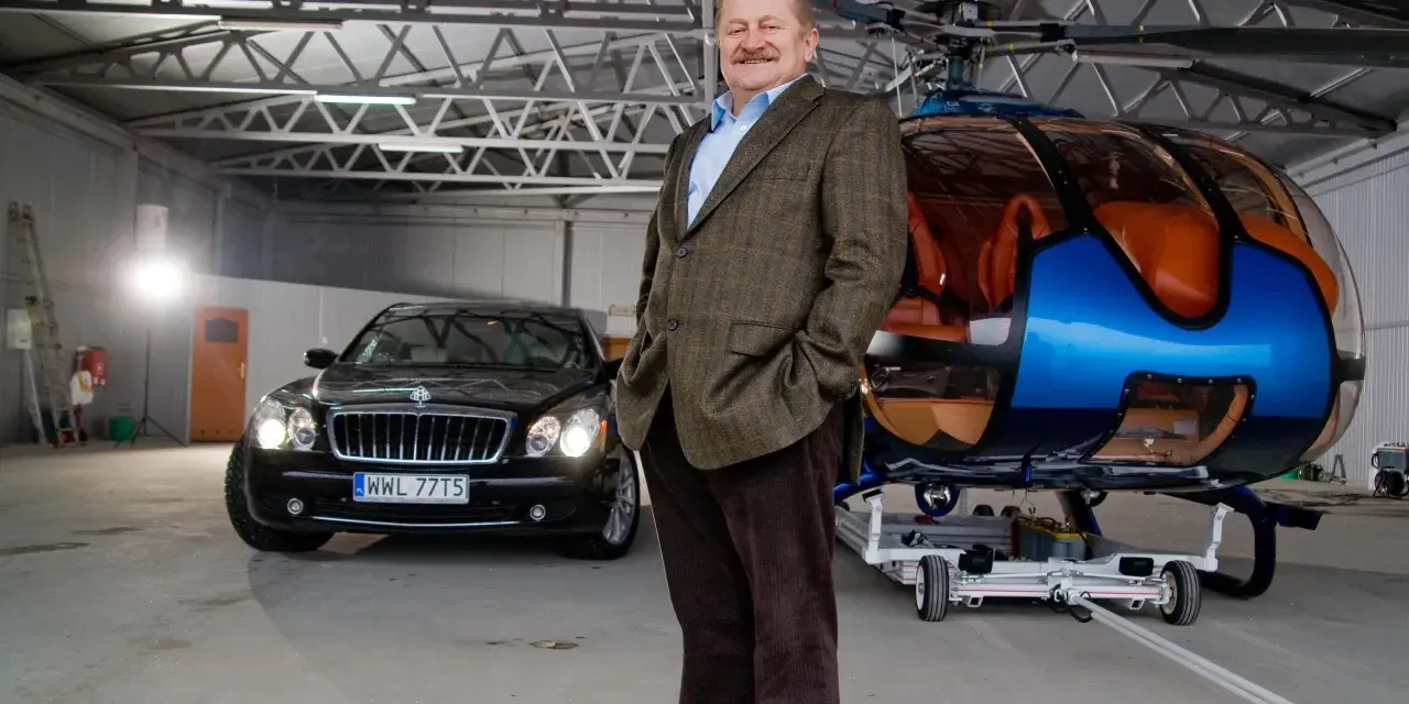 Jarosław Gołębiewski Tago w hangarze obok luksusowego samochodu i helikoptera.