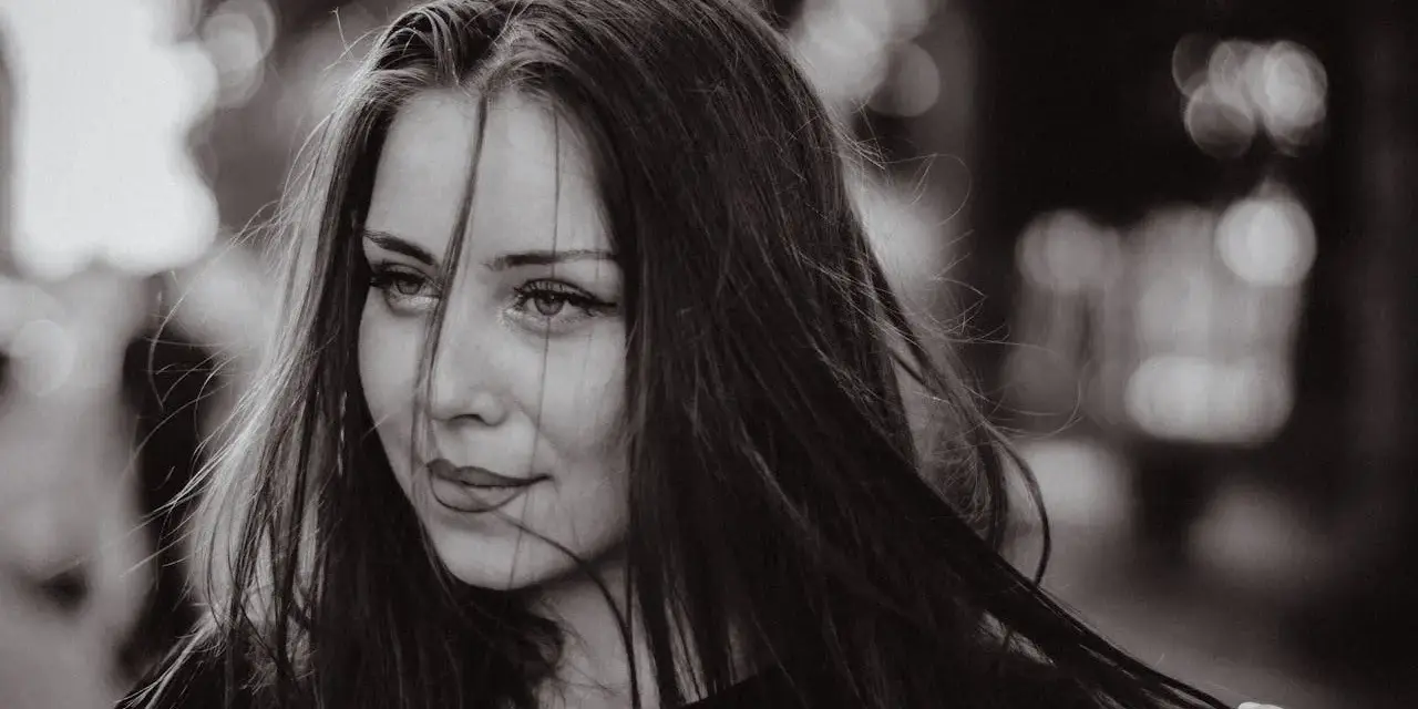 Portret młodej kobiety z burzą ciemnych włosów. Jej spojrzenie jest lekko zadumane, a usta subtelnie uśmiechnięte. Grzywka dodaje jej tajemniczości.