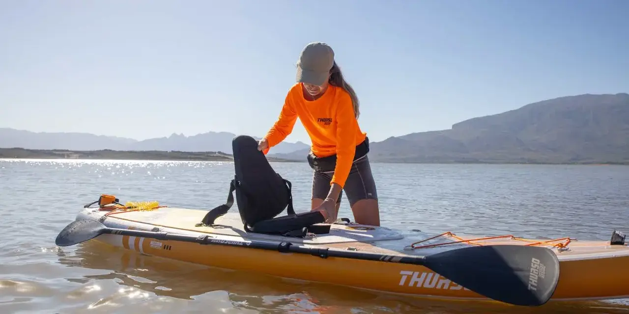 Frau richtet Sitz auf SUP-Board ein, das wie ein Kajak genutzt werden kann. Sonnenschein, Berge und See im Hintergrund.
