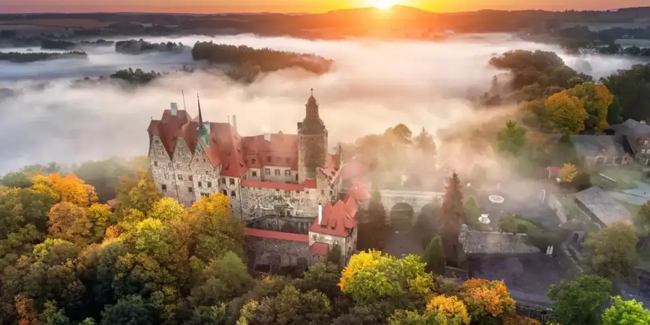 Zamek Książ otoczony jesiennymi drzewami, wyłaniający się z porannej mgły. Słońce wschodzi nad wzgórzami, malując niebo na pomarańczowo.
