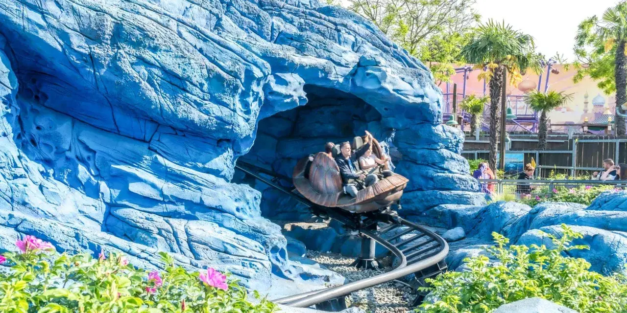 Personas en un coche de montaña rusa saliendo de una cueva azul rocosa, con palmeras y un paisaje de cuento de hadas al fondo.