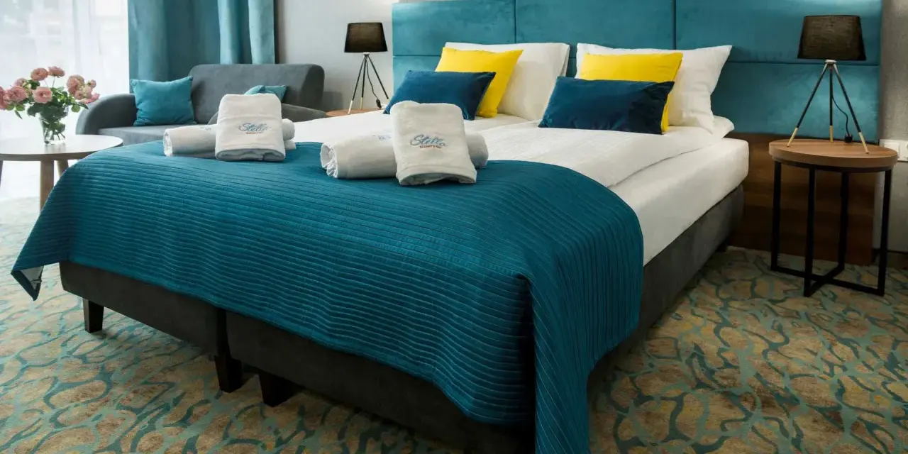 Przytulny pokój hotelowy z dużym łóżkiem, ozdobionym turkusową narzutą i żółto-niebieskimi poduszkami. Ręczniki z logo Stella Resort & Spa zachęcają do relaksu.