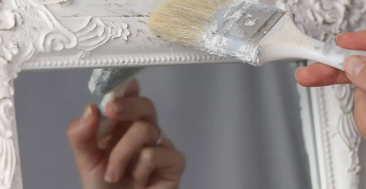 Pintura a la tiza blanco antiguo en un marco de espejo ornamentado. Una mano aplica la pintura con una brocha, creando un acabado desgastado.
