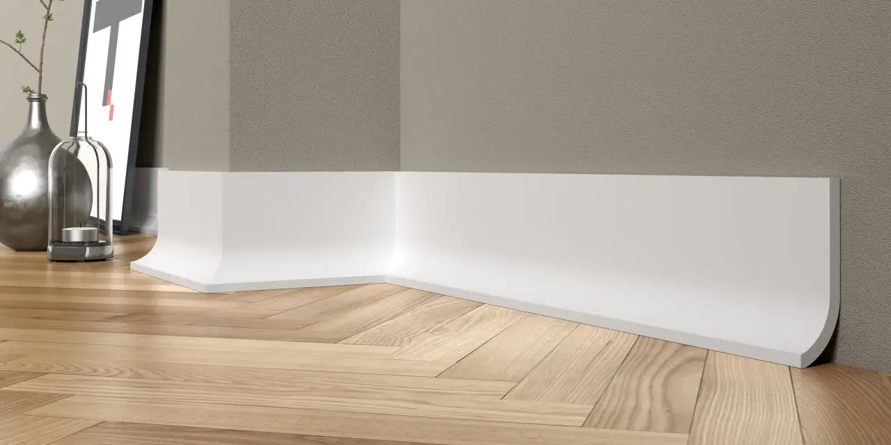 Białe, nowoczesne cokoły tworzą elegancką linię przy drewnianej podłodze w jodełkę. W tle widać fragment ściany i dekoracje.