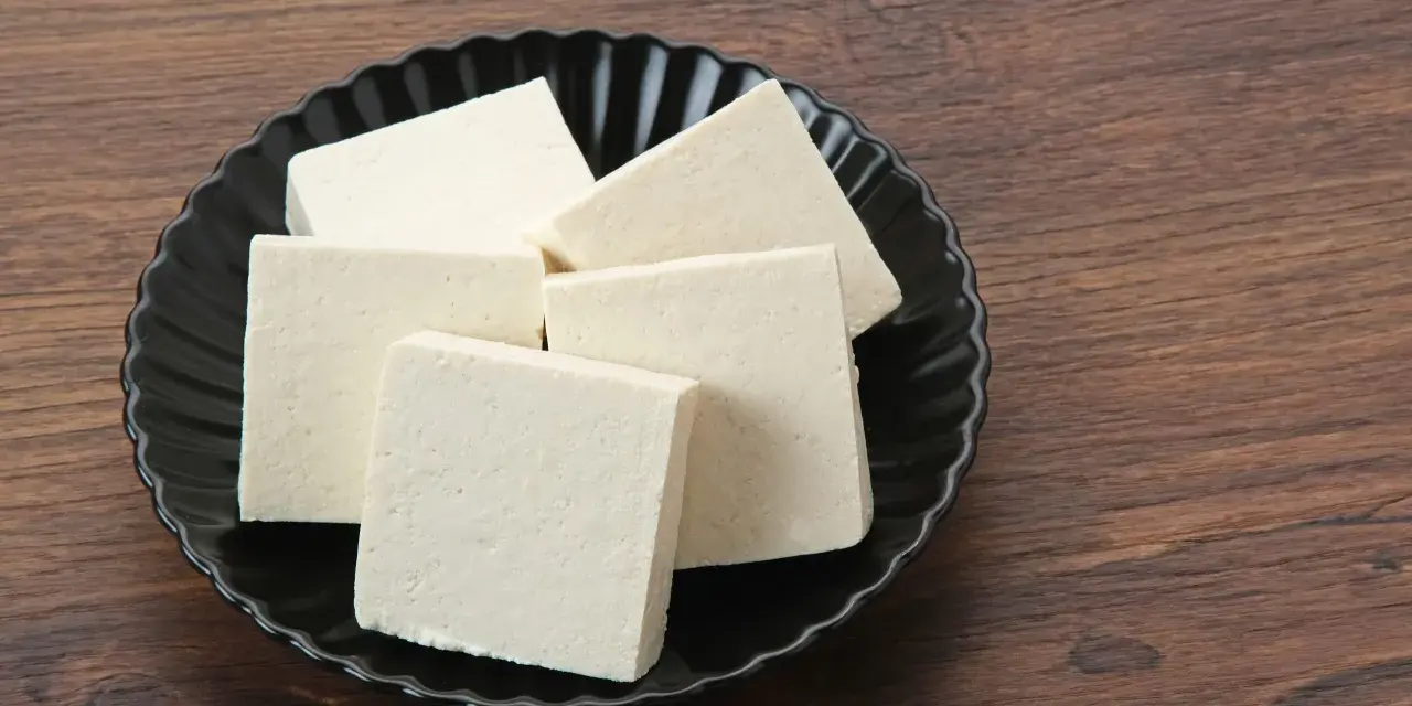 Kilka kostek białego tofu na czarnym talerzu. Czy tofu można mrozić? Tak, po rozmrożeniu zmienia teksturę, stając się bardziej gąbczaste.