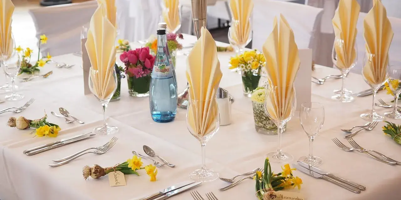 Elegancko nakryty stół weselny z żółtymi serwetkami, kwiatami i butelką wody. Każde miejsce ma imienną wizytówkę, jak dla drużby.