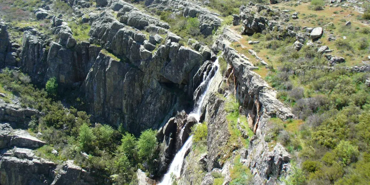 Cascada cayendo por rocas escarpadas cerca de un pico ocejon, rodeado de vegetación verde y rocas.