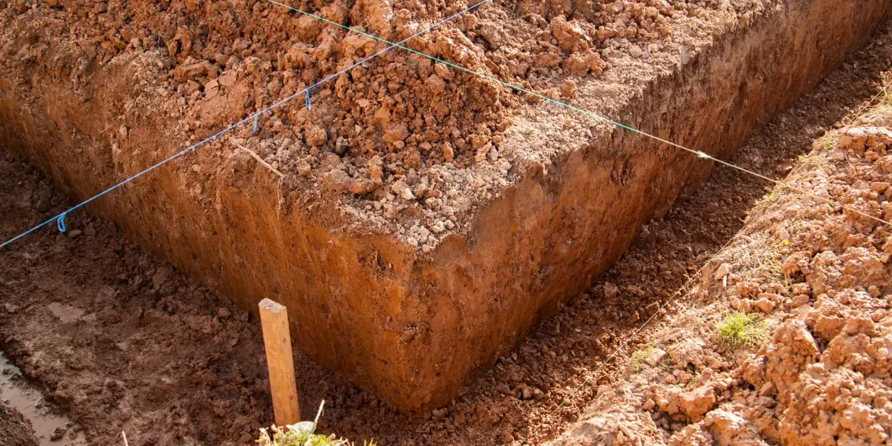 Głęboki wykop pod fundament, z widocznymi nićmi wyznaczającymi linie budowy. Widać błotniste dno i usypaną ziemię.