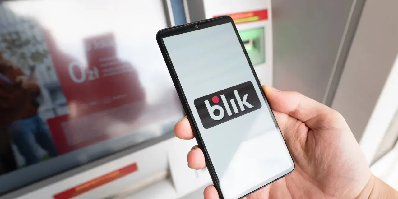 Dłoń trzyma smartfon z logo BLIK przed bankomatem.