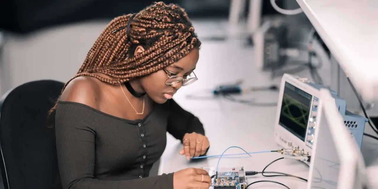 Junge Frau mit geflochtenen Haaren arbeitet konzentriert an einer Platine, während ein Oszilloskop im Hintergrund läuft. Ein spannendes elektrotechnik studium.
