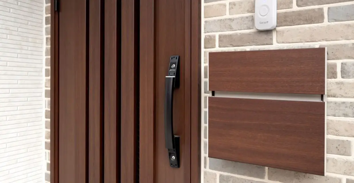Puerta de madera oscura con tirador negro, buzón de madera y un timbre blanco. Varios tipos de timbre se pueden ver en la pared de ladrillos.